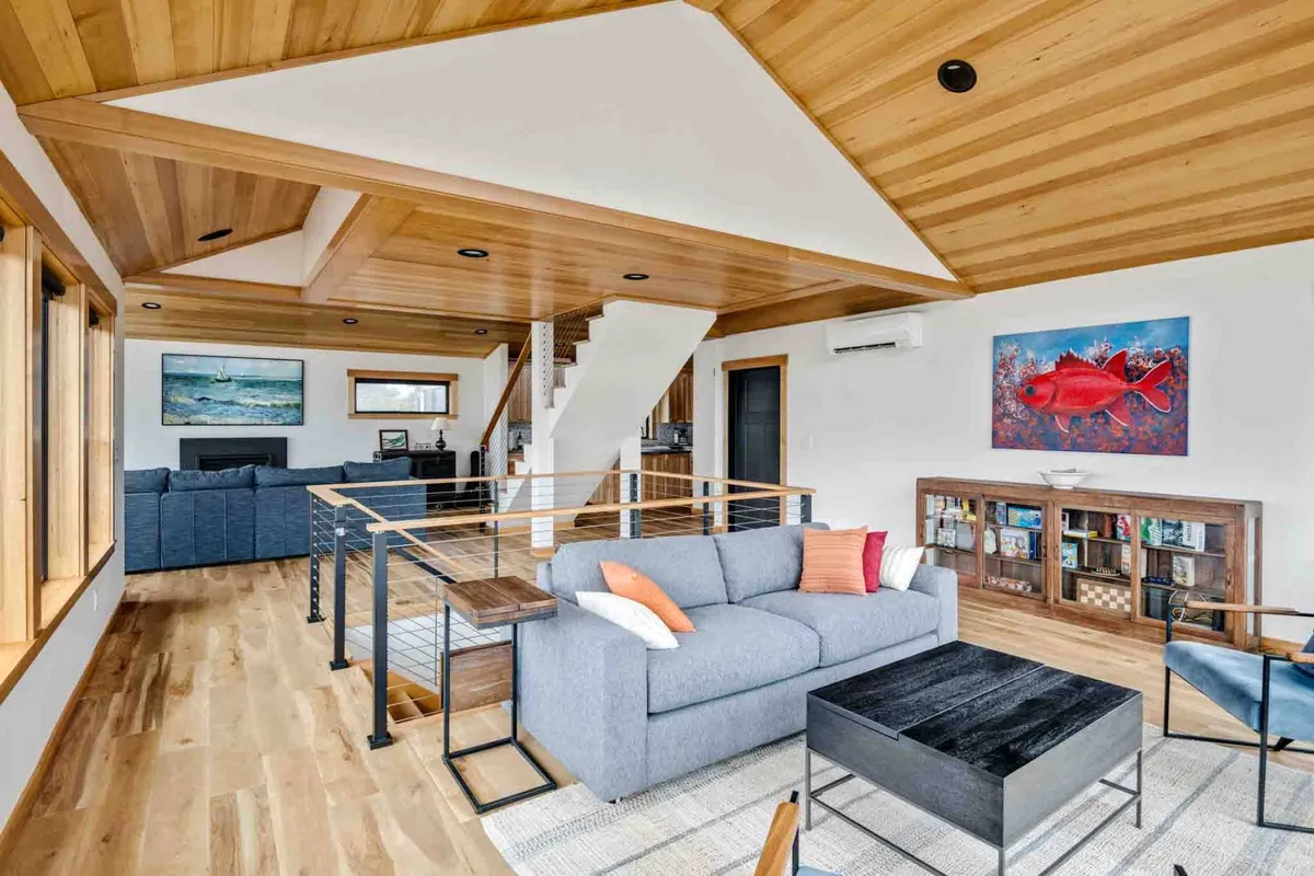 A stylish living room featuring a comfortable sofa with colorful cushions, eye-catching fish artwork, wooden railings, and a stairway leading to the upper floor. The space is inviting and hints at extra sleeping flexibility for groups or families.