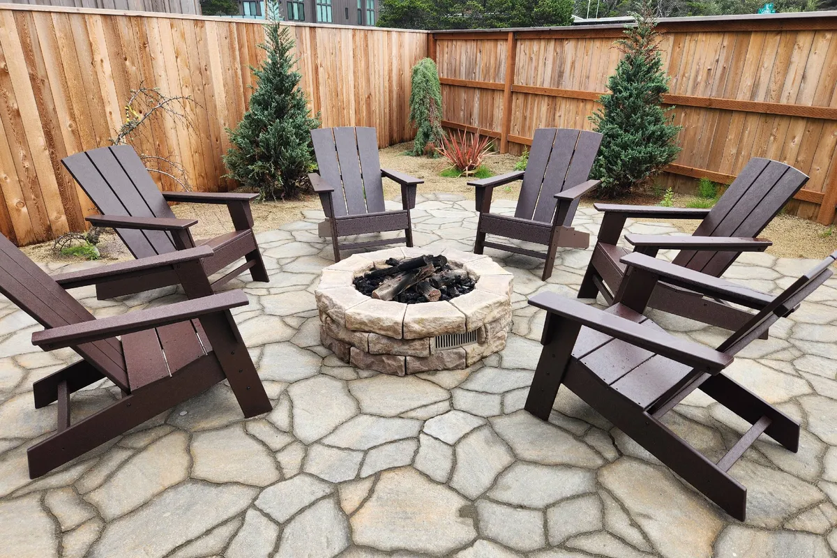 A cozy communal firepit area is shown with Adirondack chairs arranged in a circle on a stone patio, surrounded by a wooden fence and small shrubs. The setting evokes warmth, community, and evenings spent sharing stories under the stars.
