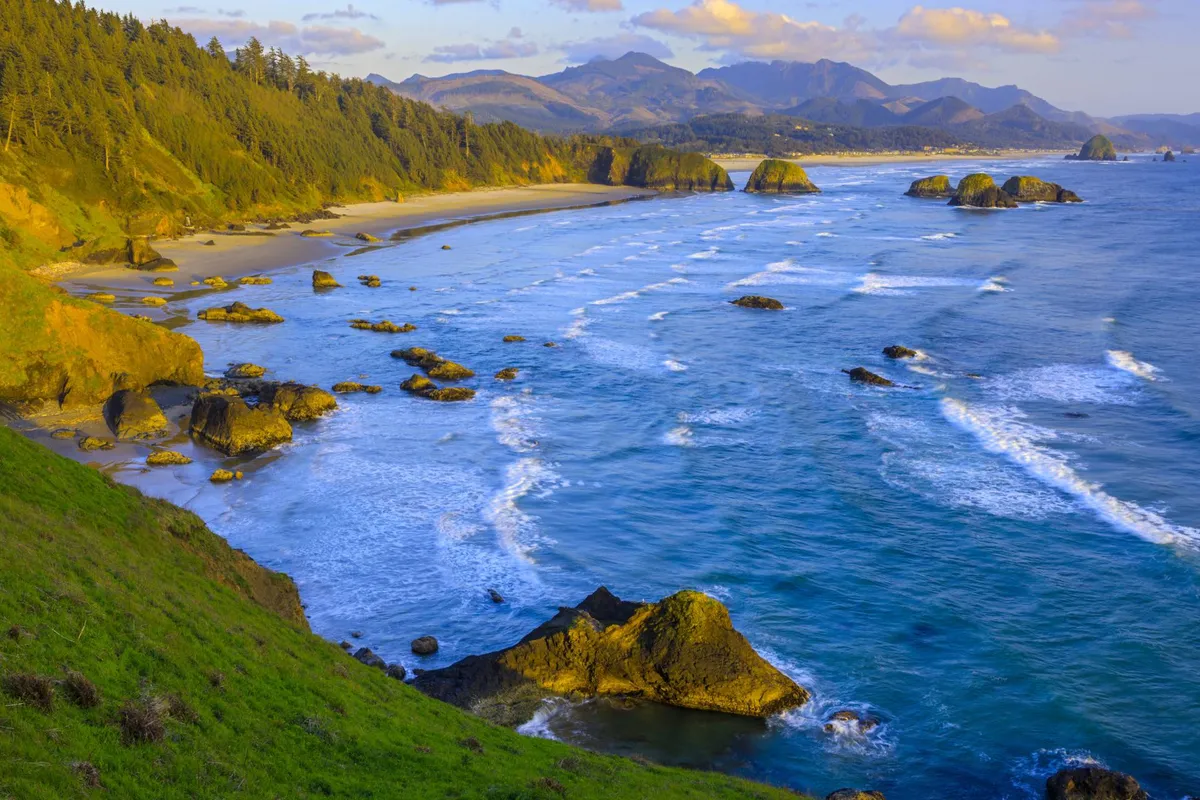 A stunning view of the rocky coastline, gentle waves, lush greenery, and distant mountains. The natural beauty and open coastal landscape invite exploration and relaxation, aligning with the activities like beachcombing and photography.