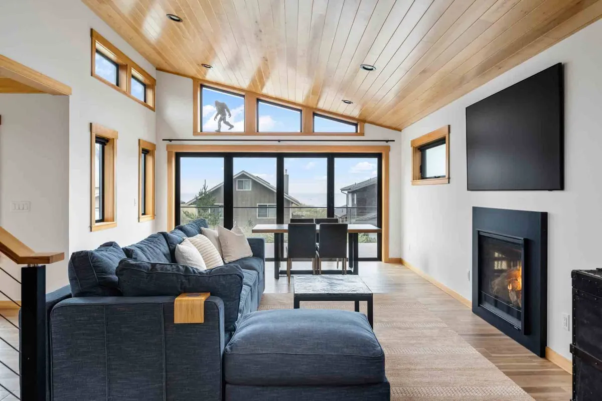 A spacious living area featuring a large, comfortable blue sectional sofa arranged around a modern fireplace, with expansive windows letting in abundant natural light and offering scenic views. The room also includes a dining table with chairs and striking wooden ceiling details, creating an inviting open-concept layout.