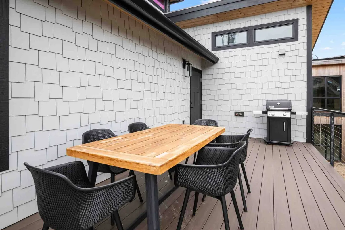 An inviting outdoor dining area featuring a wooden table surrounded by black mesh chairs, with a barbecue grill nearby. The patterned wall and open-air setup suggest a perfect spot for evening meals and social gatherings as the sun sets.