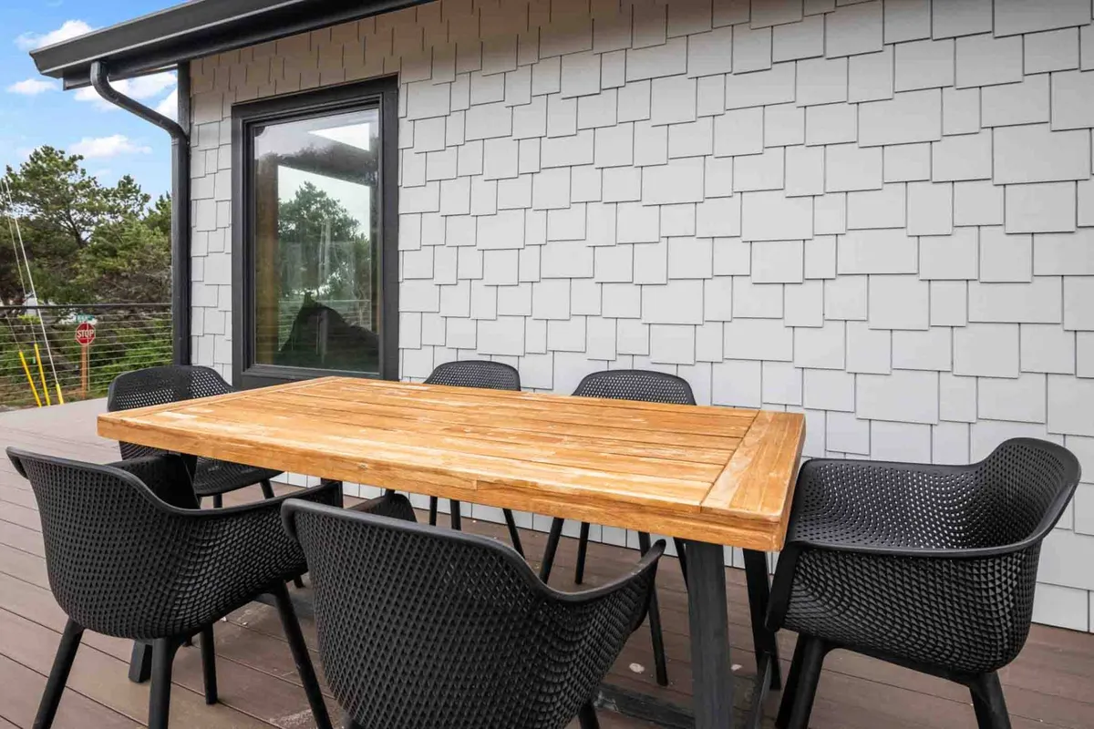 A cozy exterior patio space featuring a wooden dining table and black chairs, set against a textured wall and framed by large glass windows. The area is ideal for outdoor dining, relaxing, or gathering with friends while enjoying the fresh coastal air.