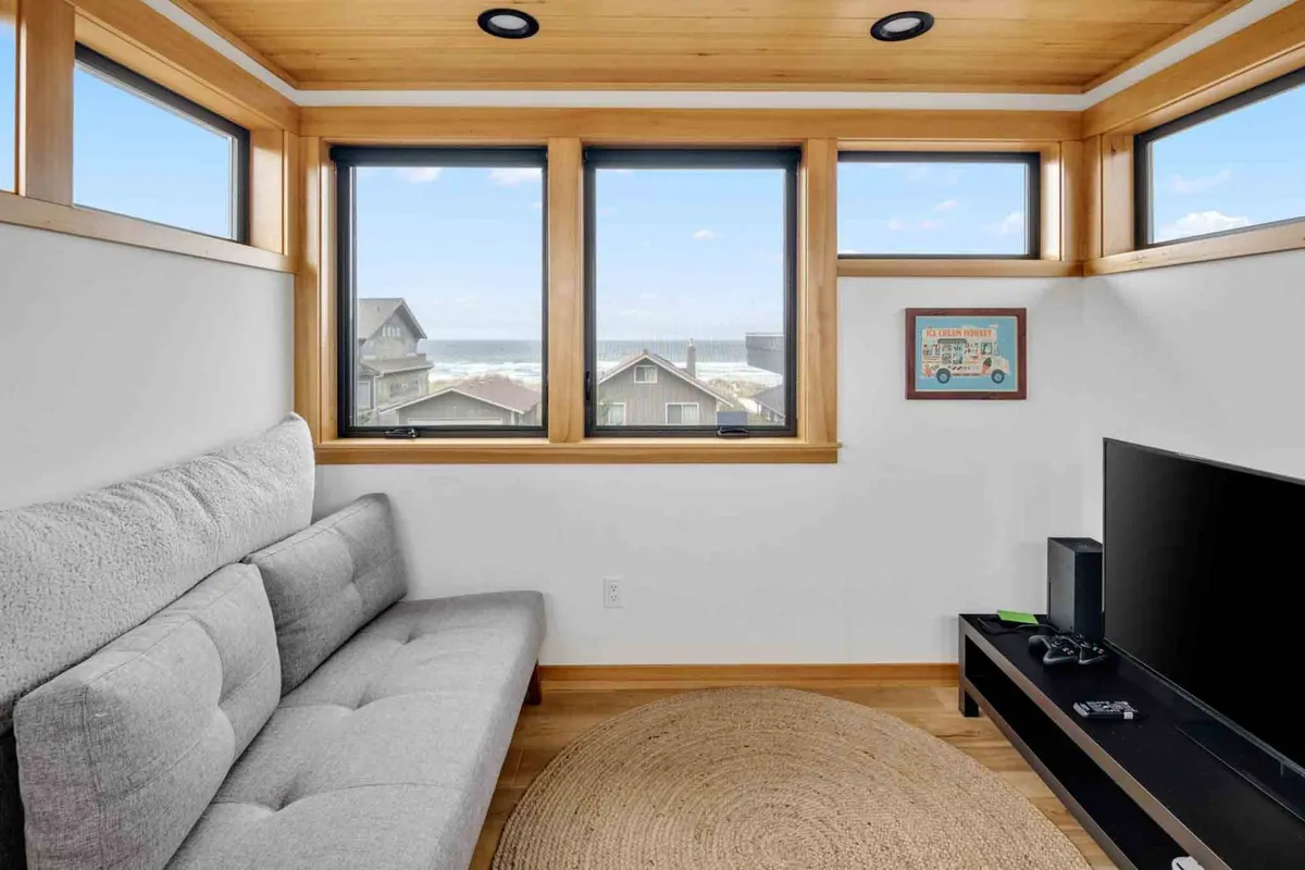 A luminous living room with large windows offering an expansive view, a modern sofa, a TV unit with a gaming console, and decorative artwork. The room is designed for relaxing, socializing, and enjoying panoramic scenes, possibly of the ocean or surrounding landscape.
