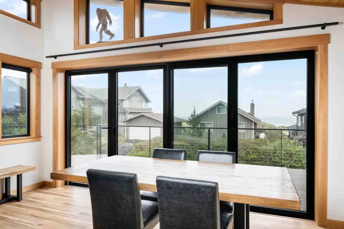A welcoming dining area with a large table set by expansive glass windows, offering views of surrounding houses and the evening sky. The scene suggests warmth, comfort, and togetherness during family meals or game nights.