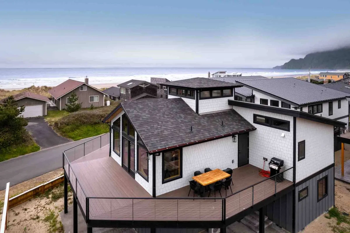The image showcases the sleek, modern exterior of the 'Vitamin Sea' beach house in Manzanita, Oregon. The spacious deck is outfitted with a dining table perfect for outdoor meals, and the scene captures expansive ocean views, hinting at stunning sunsets and the relaxing ambiance of the coast. The setup suggests opportunities for alfresco dining, barbecues, and family gatherings, all while enjoying the fresh sea breeze.