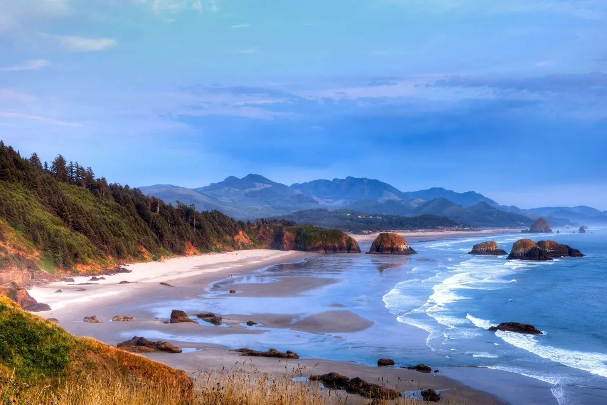 A sweeping view of the Oregon Coast featuring dramatic rock formations, a wide sandy beach, rolling waves, and tree-lined mountains in the distance. The tranquil setting invites outdoor pursuits such as hiking, beachcombing, and wildlife observation.