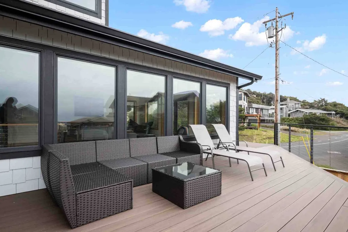 A spacious outdoor seating area on the deck features a glass table surrounded by comfortable chairs, offering unobstructed views of the surrounding landscape. The area is bathed in natural light and appears perfectly suited for relaxing, entertaining, or enjoying meals outside while soaking in coastal scenery.